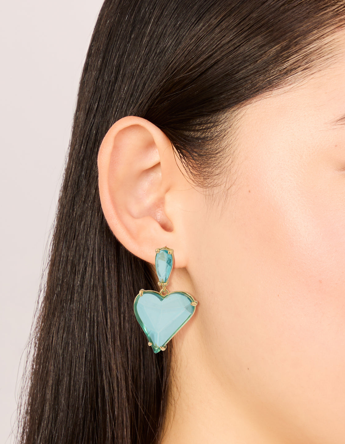 Displaying statement drop earrings with heart design in blue glass stone and gold finish on ear view